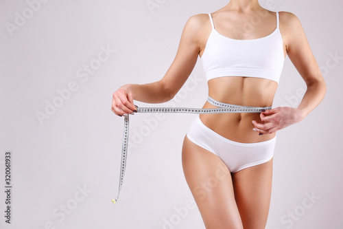 Close up shot of unrecognizable fit woman in lingerie measuring her waist with a tape isolated on white background. Torso of slim attractive female with flat belly in white underwear. Copy space.