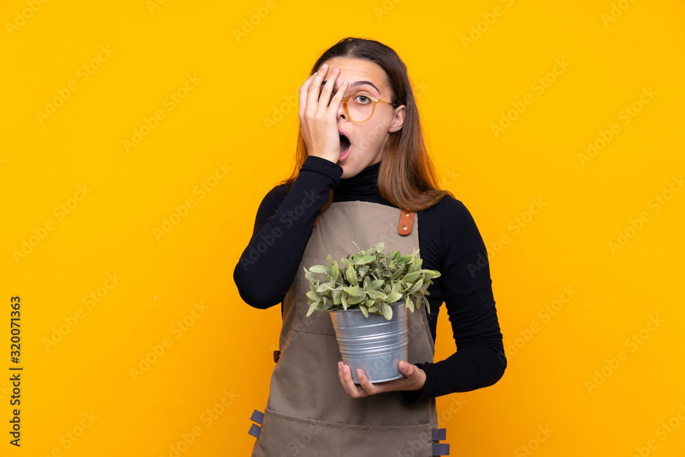 Young gardener girl holding a plant over isolated yellow background covering eyes and looking through fingers Young gardener girl holding a plant over isolated yellow background covering eyes and looking through fingers