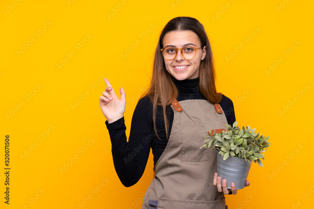 Young gardener girl holding a plant over isolated yellow background pointing finger to the side Young gardener girl holding a plant over isolated yellow background pointing finger to the side