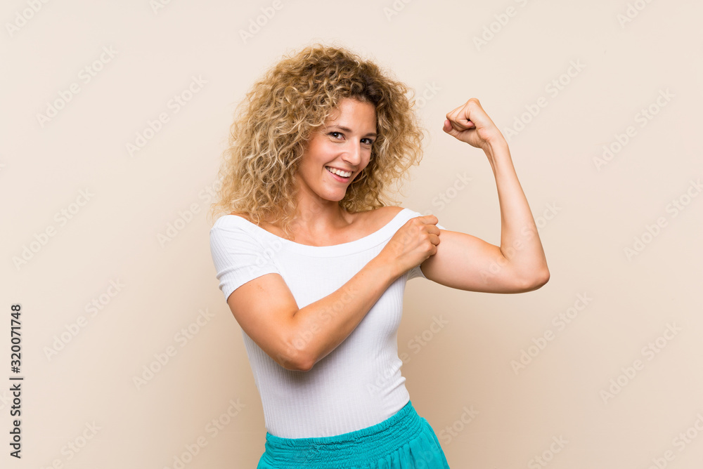Young blonde woman with curly hair over isolated background doing strong gesture Young blonde woman with curly hair over isolated background doing strong gesture