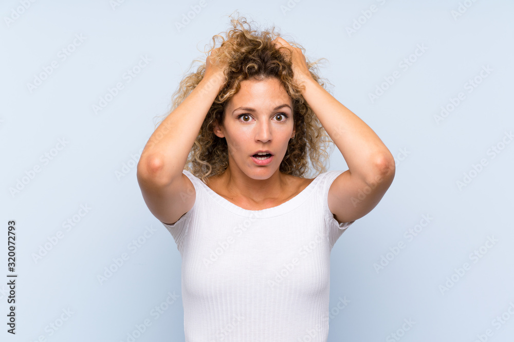 Young blonde woman with curly hair over isolated blue background with surprise facial expression Young blonde woman with curly hair over isolated blue background with surprise facial expression