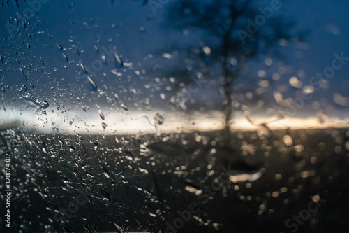 Raindrops on the side window of the car against the background of the sunset and the silhouette of a tree, a photo from inside the car