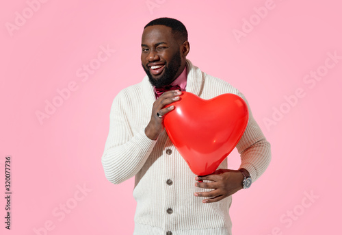 Handsome man winking. Photo of african american man in love holds red heart shape balloon on pink background © Romario Ien