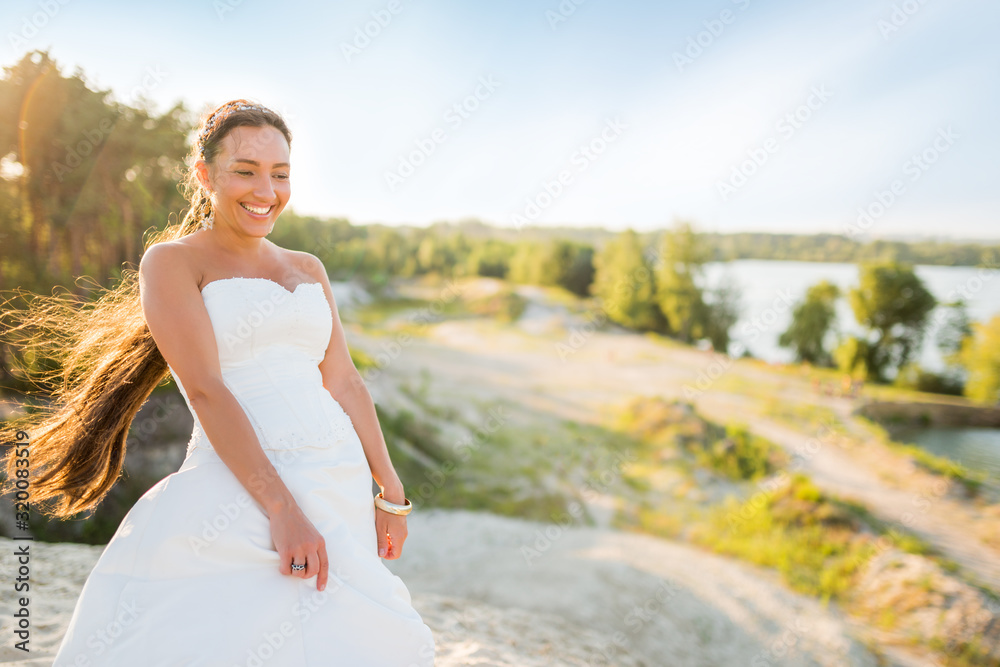 Beautiful young woman bride posing on white sand against the backdrop of a pine forest on a sunny warm summer day. Romantic wedding concept