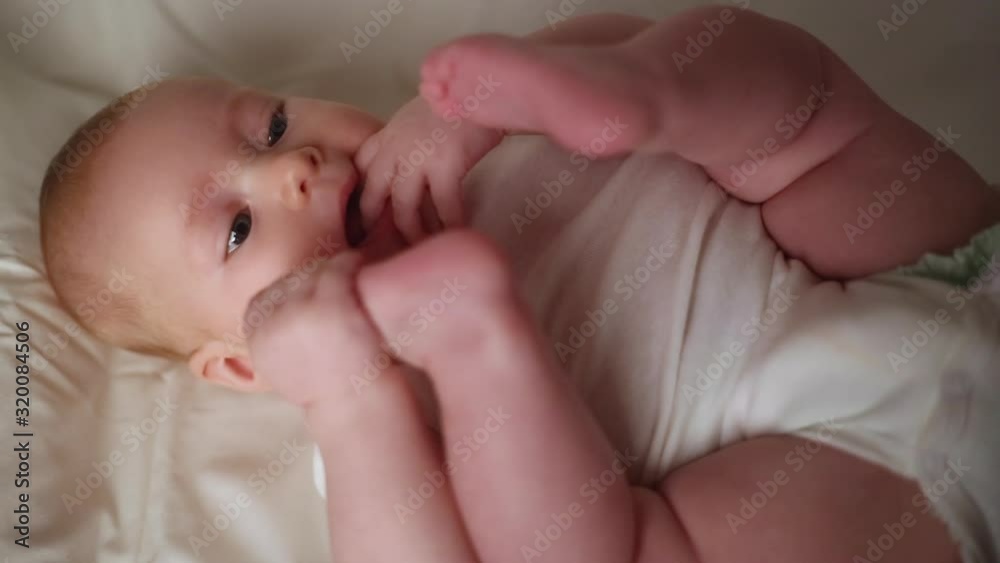 The child is trying to eat his legs. Little cute girl lies on her back in a white bodysuit and puts her feet in her mouth. The child is trying to eat his legs. Little cute girl lies on her back in a white bodysuit and puts her feet in her mouth.