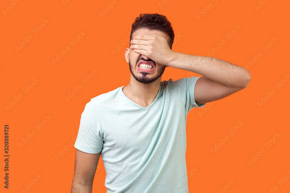 Don't want to look at this. Portrait of depressed brunette man with beard in white t-shirt covering eyes, ignoring problems with angry scared face. indoor studio shot isolated on orange background Don't want to look at this. Portrait of depressed brunette man with beard in white t-shirt covering eyes, ignoring problems with angry scared face. indoor studio shot isolated on orange background