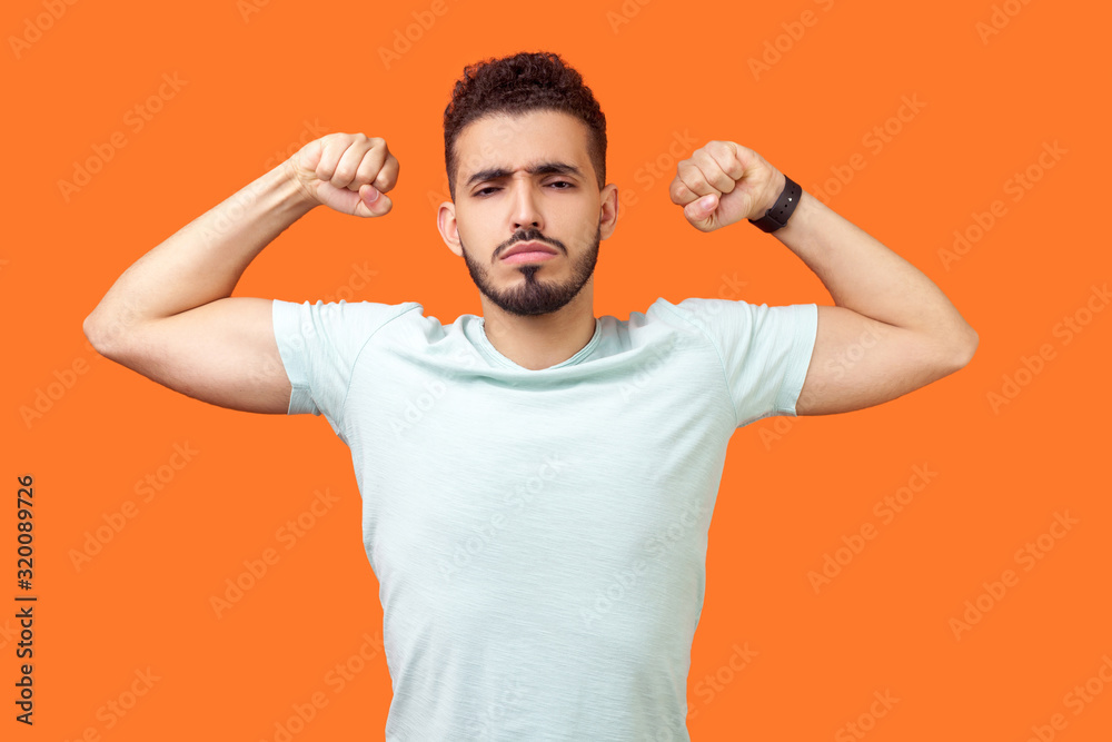 I am strong and powerful. Portrait of confident successful brunette man with beard in white t-shirt showing biceps, full of motivation and willpower. indoor studio shot isolated on orange background I am strong and powerful. Portrait of confident successful brunette man with beard in white t-shirt showing biceps, full of motivation and willpower. indoor studio shot isolated on orange background