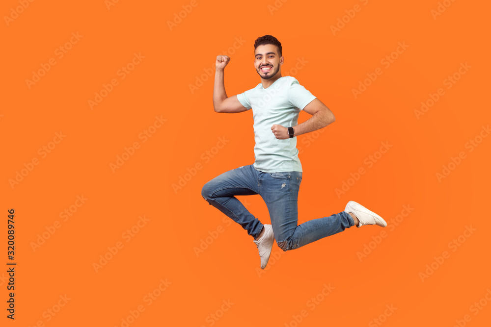 Full length portrait of positive inspired brunette man with beard in sneakers, denim outfit jumping in air or running quickly fast. indoor studio shot isolated on orange background, empty copy space