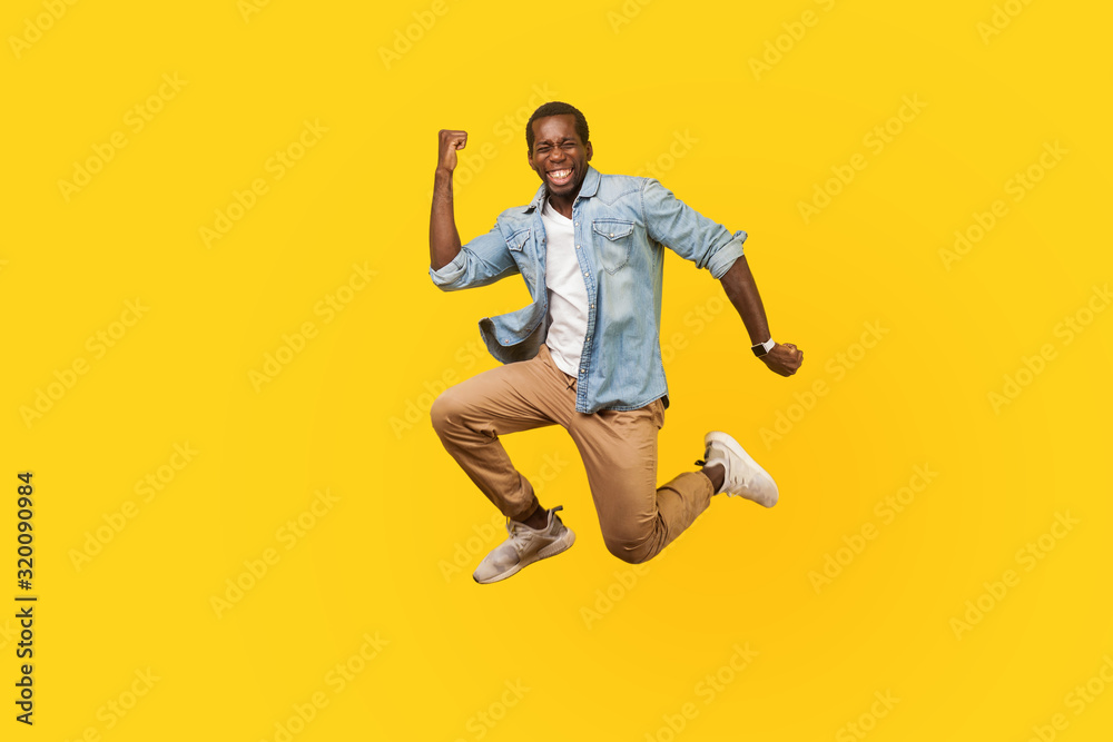 Full length portrait of joyous ecstatic man in denim shirt jumping for joy or flying with raised hand, gesturing yes i did it, celebrating success. indoor studio shot isolated on yellow background