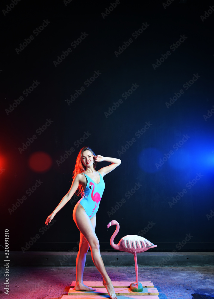 Fototapeta premium beautiful sexy girl with red hair in a swimsuit with a flamingo pattern posing with a pink flamingo toy in colored smoke
