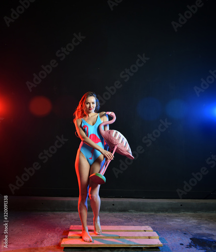 Fototapeta Naklejka Na Ścianę i Meble -  beautiful sexy girl with red hair in a swimsuit with a flamingo pattern posing with a pink flamingo toy in colored smoke
