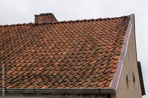 Fragment of old  worn brown tile roof on the right of house with brick chimney   beige-plastered gable with small windows on it and a metal storm drain running horizontally under the edge of the roof
