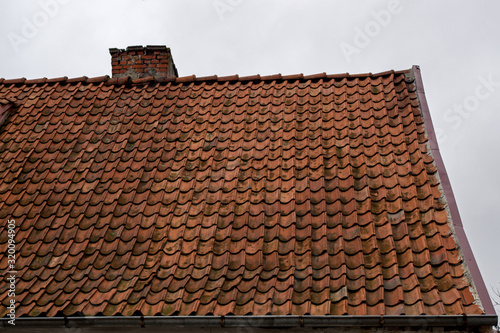 A fragment of an old shabby brown beige gable tiled roof of an old house with a brick chimney stack furnace with a rain gutter under it against the background of a gloomy gray overcast sky © ANDY RELY