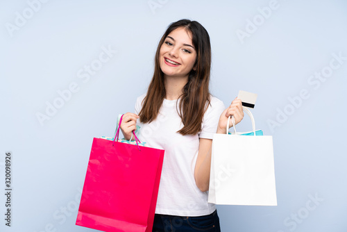 Young brunette woman over isolated blue background holding shopping bags and a credit card