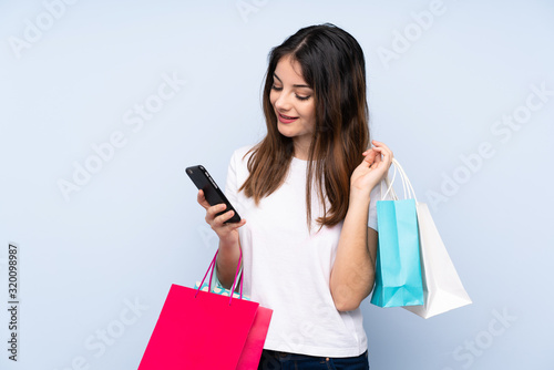 Young brunette woman over isolated blue background holding shopping bags and writing a message with her cell phone to a friend