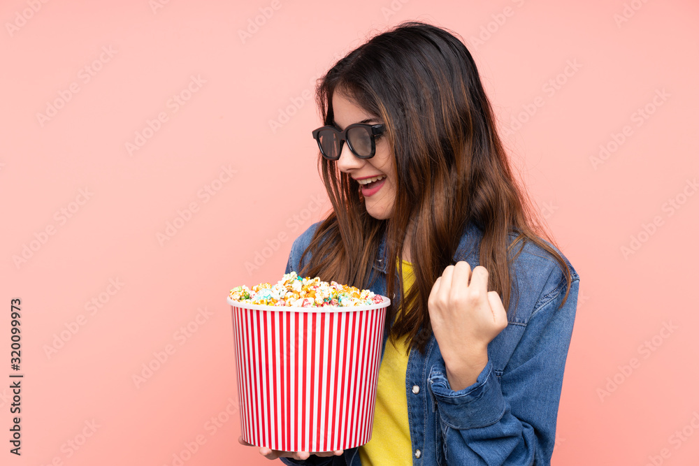 Young brunette woman over isolated pink background with 3d glasses and holding a big bucket of popcorns while looking side