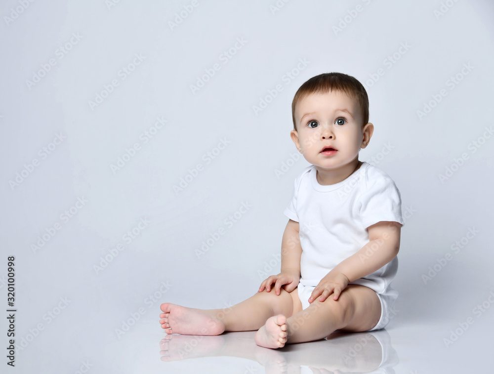 Infant baby boy toddler in white bodysuit is sitting sideways on the floor looking up at us on a gray with copy space