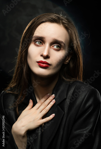 Beautiful slim girl model with red lips  wearing a black blazer  with an apologetic gesture presses her hand to her chest on a dark background  next to the light lamp. Advertising  trendy design.