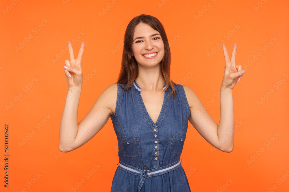Peace and victory to you! Friendly cheerful brunette woman in denim dress making double v gesture and smiling at camera, showing sign of success, triumph. studio shot isolated on orange background Peace and victory to you! Friendly cheerful brunette woman in denim dress making double v gesture and smiling at camera, showing sign of success, triumph. studio shot isolated on orange background