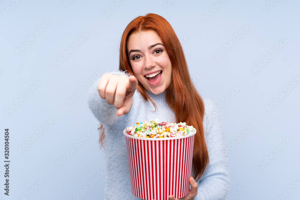 Redhead teenager girl over isolated blue background holding a big bucket of popcorns while pointing front