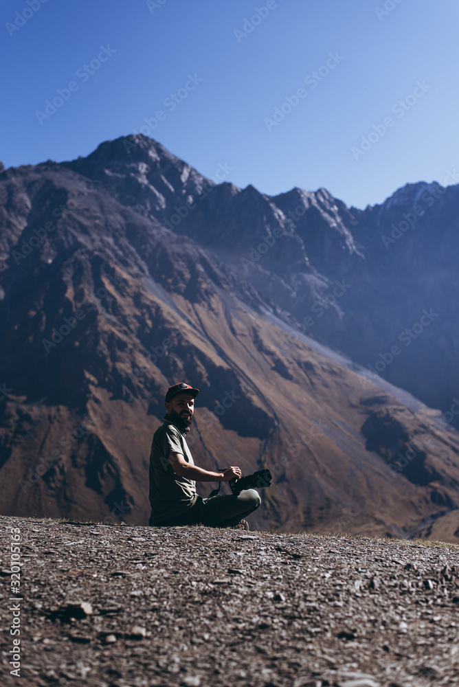 Fototapeta premium stylish bearded man with camera in hands on top of Georgian mountains: nature photographer traveler taking photo of beautiful landscape from top of the mountain