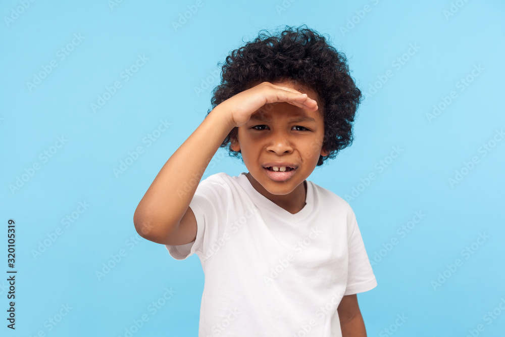 Portrait of astonished little boy with curls holding hand over eyes and looking attentively far away, trying to see long distance, expressing disbelief and amazement. studio shot, blue background - obrazy, fototapety, plakaty 