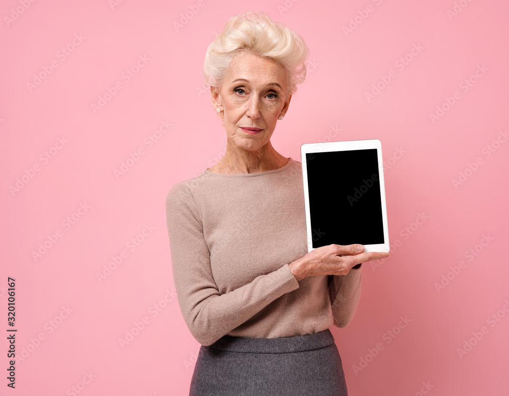 Smiling woman holding a digital tablet with a blank screen. Photo of elderly woman on pink background.