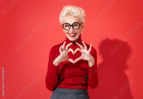 Beautiful woman shows heart symbol, shapes love sign with hands. Photo of smiling elderly woman in love on red background. Be my Valentine