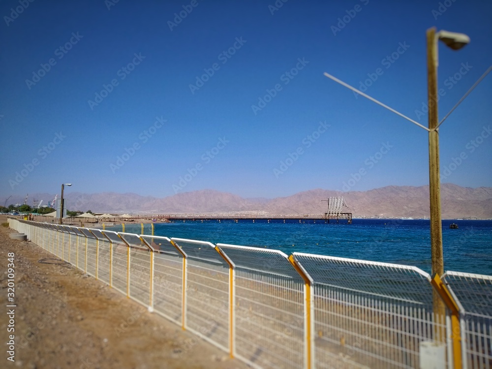 shiny fence. In the background is an empty beach and a turquoise ocean. White clouds on a blue sky