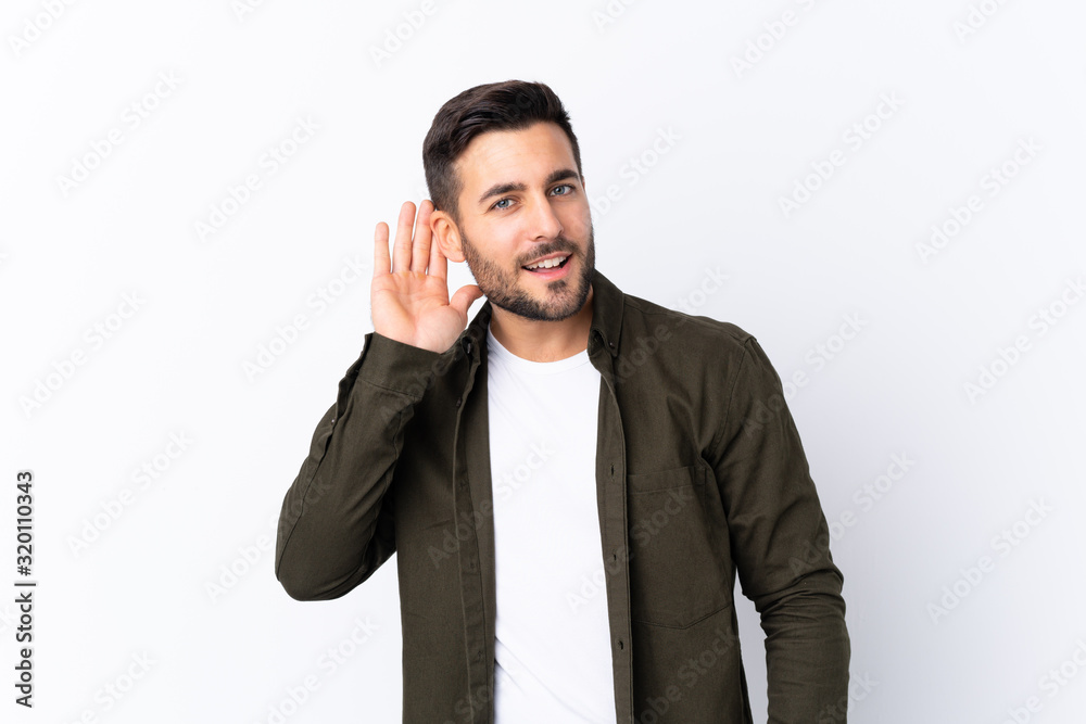 Young handsome man with beard over isolated white background listening to something by putting hand on the ear Young handsome man with beard over isolated white background listening to something by putting hand on the ear