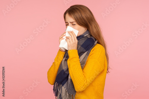 Flu season. Portrait of unhealthy red hair girl wearing big scarf, sneezing and blowing nose, wiping with tissue, feeling unwell, influenza symptoms. indoor studio shot isolated on pink background