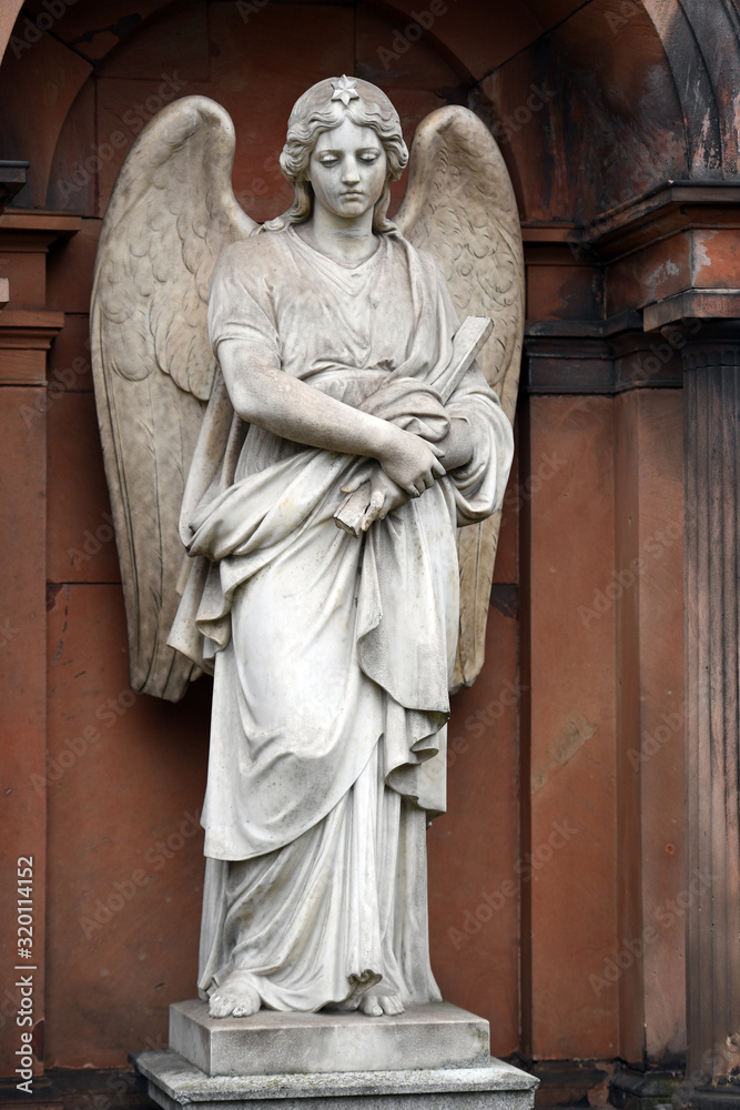 A weathered, white sandstone sculpture of a grieving angel with spread wings in front of an old crypt of an cemetery in Berlin-Germany.