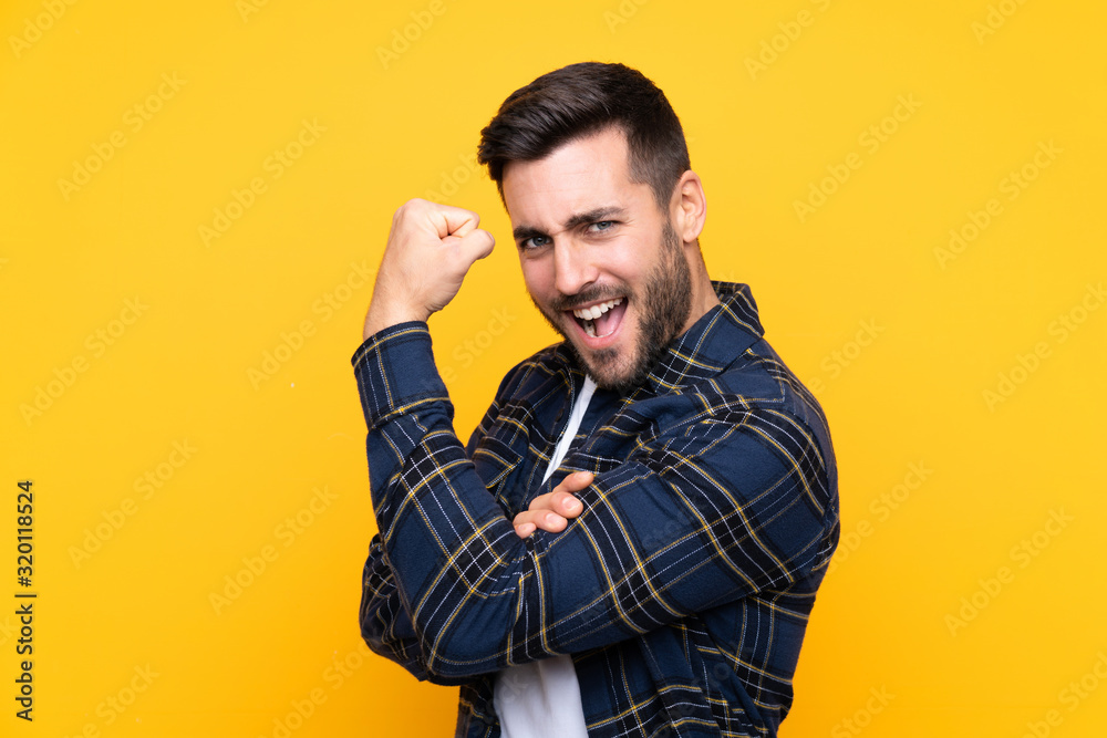 Young handsome man with beard over isolated yellow background doing strong gesture Young handsome man with beard over isolated yellow background doing strong gesture
