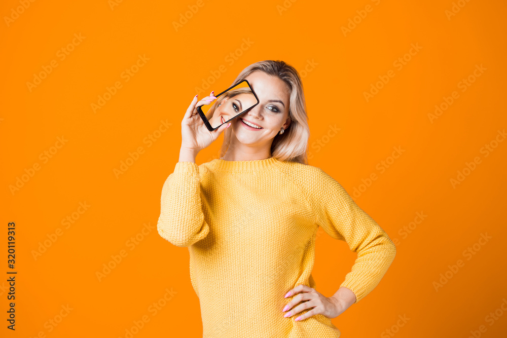 young blonde in a yellow sweater holds a smartphone in her hand with her selfie on the screen.