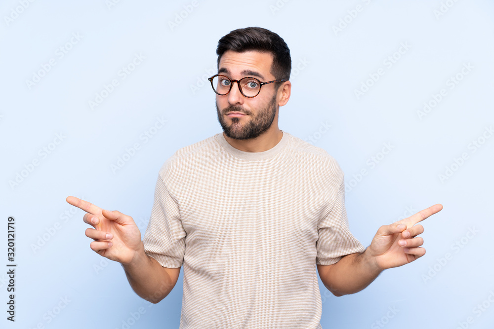 Young handsome man with beard over isolated blue background pointing to the laterals having doubts Young handsome man with beard over isolated blue background pointing to the laterals having doubts