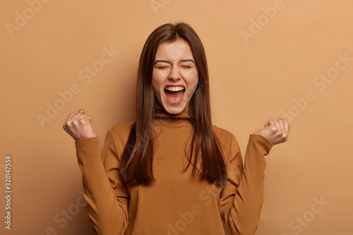 Overjoyed brunette woman make fists pump, celebrates success, shouts and feels like champion, keeps mouth wide opened, wears turtleneck, isolated over beige wall, exclaims hooray finally I did it photo