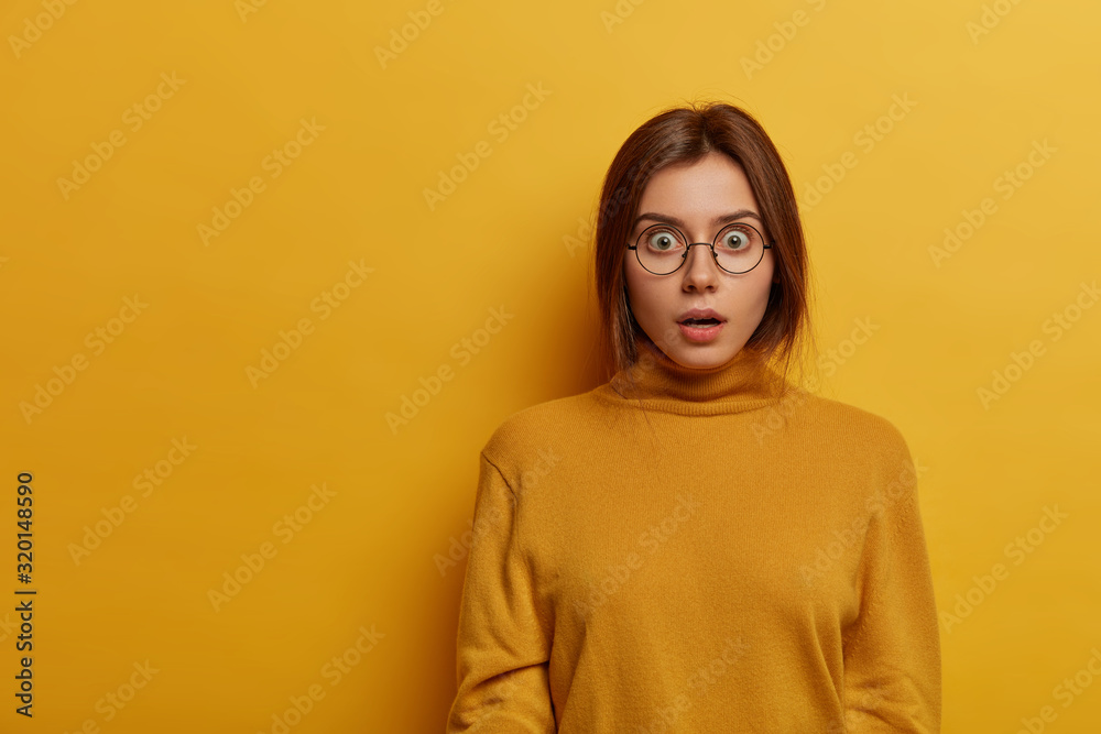 Stunned emotional woman lost much money, stares speechless and concerned, nervously reacts on terrible situation, wears round big glasses and casual sweatshirt, isolated over yellow background Stunned emotional woman lost much money, stares speechless and concerned, nervously reacts on terrible situation, wears round big glasses and casual sweatshirt, isolated over yellow background