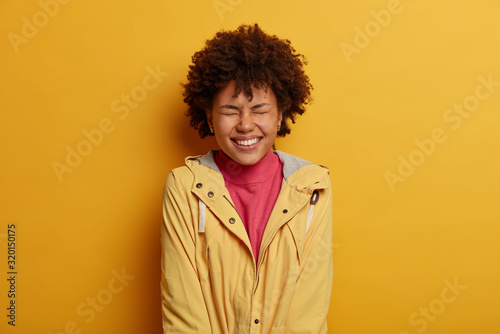 Overemotive cheerful woman closes eyes and smiles broadly, feels very happy after successful day, cannot wait outstanding event, wears windbreaker, shows white teeth, isolated on yellow studio wall