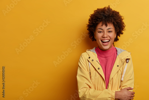 Enthusiastic hilarious woman with Afro hairstyle, laughs out loud, imagines funny situation, keeps hands crossed over chest, dressed casually, stands against yellow wall, copy space, smiles happily