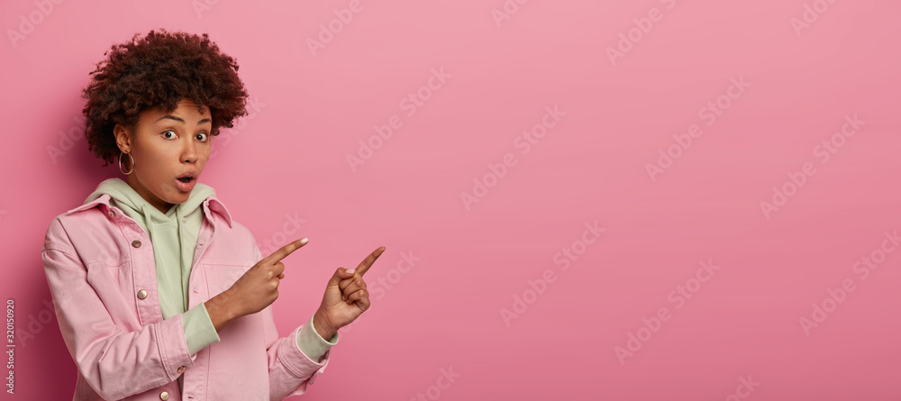 Amazed stunned dark skinned woman points index fingers on copy space area, looks with puzzlement and confusion, opens mouth, dressed in casual jacket, isolated on pink studio wall. Omg, look there - obrazy, fototapety, plakaty 