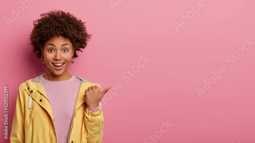 Positive curly haired woman points thumb aside, has glad expression, demonstrates something interesting, wears casual jacket, poses over rosy wall with empty space. People and advertisement. photo