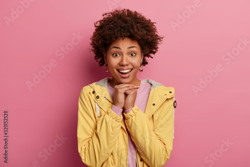 Portrait of beautiful positive woman smiles gladfully, keeps hands under chin, looks directly at camera, looks forward to exciting event, awaits for surprise, wears yellow anorak, models indoor