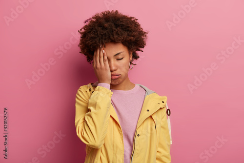 Tired female teenager sighs from unhappiness, covers face with palm, closes eyes and suffers from headache, being overworked, has sleepy expression wears casual anorak, stand over rosy studio wall.