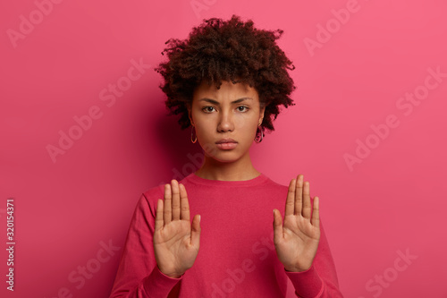 No means never. Strict serious looking woman pulls palms forward at camera in no gesture, rejects offer, asks to stop, warns about danger, wears crimson jumper, says calm down, control behaviour