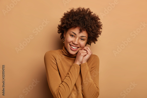 People and pleasant emotions concept. Gentle positive woman tilts head, smiles with delight, gazes tenderly at camera, feels happiness, wears casual jumper, has cheerful grin, isolated on beige wall