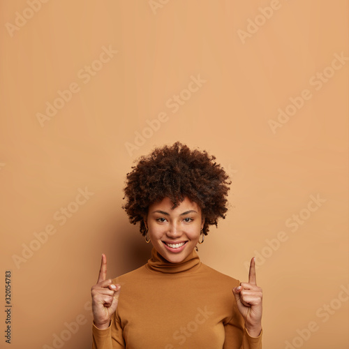 Cheerful curly woman keeps index finger pointed upward, shows something above head, smiles positively, makes advertising gesture, wears casual turtleneck, gives good advice, space for your text