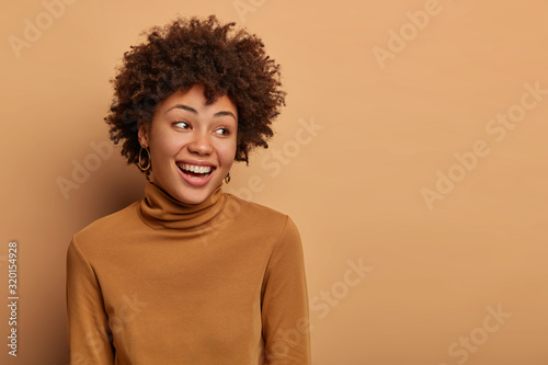 Half length shot of tender female looks gladfully aside, smiles sincerely, has natural curly hair, being in high spirit, wears brown turtleneck, stands over beige wall, copy space, notices funny thing