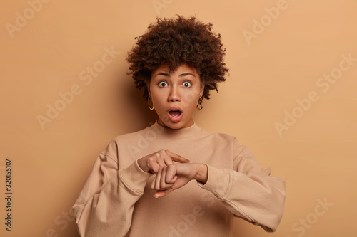 Frightened woman worries about deadline, makes time gesture, being late for important meeting, drops jaw and has bugged eyes, wears sweatshirt, isolated over brown background. Omg, be hurry! © wayhome.studio 