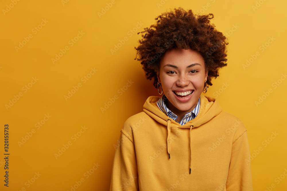 Studio shot of joyful teenager laughs sincerely, wears casual hoodie, expresses positive emotions, has casual funny talk with interlocutor, stands over yellow wall, copy space left for your text