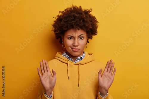 Lets calm down. Serious curly woman keeps palms outstretches, makes refusal gesture, raises hands in stop motion, rejects offer, denies something, wears yellow hoodie stands indoor unwills participate photo